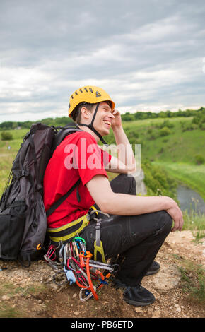 Photo of smiling smiling male tourist in yellow helmet with rucksack and carbines on hill Stock Photo