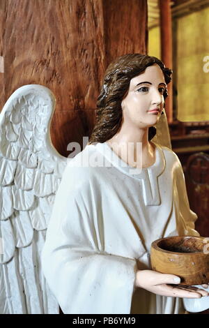 White dressed-white winged Angel statue supporting with his hands the holy water stoup at the beginning of the main nave in the Santa Monica Parish Ch Stock Photo