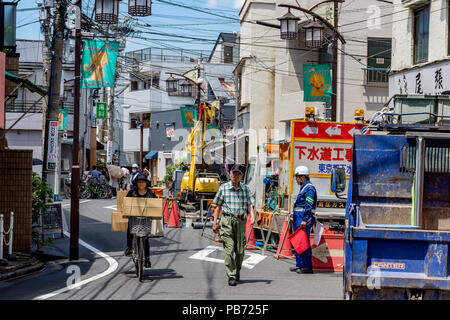 Woman on bicycle with large boxes cycles past old man walking past road construction site with guard holding red flags, in Tokyo, Japan. Stock Photo
