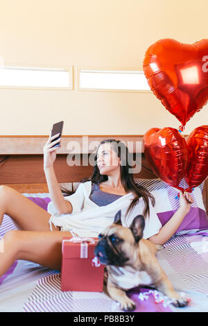 Attractive young woman laying on bed with her french bulldog and taking selfie photo. Stock Photo