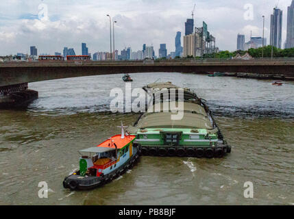 Bangkok, Thailand - May 1, 2018: Traction boat pushing a transportation boat over a river with Bangkok skyline in the background Stock Photo