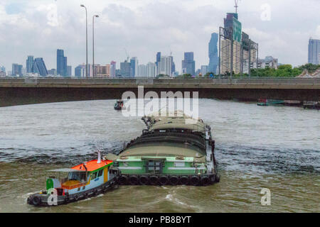 Bangkok, Thailand - May 1, 2018: Traction boat pushing a transportation boat over a river with Bangkok skyline in the background Stock Photo