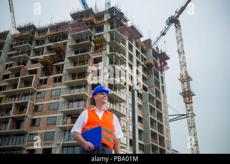 Smiling engineer holding documents on a construction site with buildings and cranes in the background Stock Photo