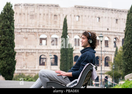 Handsome young man in sports cloths listening to music sitting on a bench with Colosseum in the background. Young sportsman in tracksuit relaxing afte Stock Photo