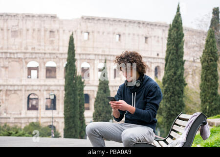 Handsome young man in sports cloths using his mobile phone while sitting on a bench in Rome. Young sportsman in tracksuit relaxing after training with Stock Photo