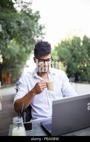 Young attractive afro American businessman sitting in cafe bar, drinking coffee and doing some work on laptop. Stock Photo