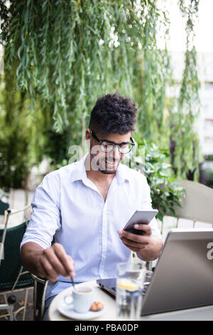 Young attractive afro American businessman sitting in cafe bar, drinking coffee and doing some work on laptop. Stock Photo