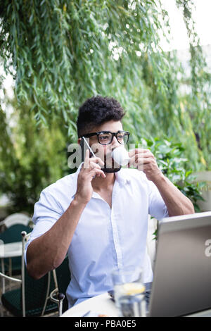 Young attractive afro American businessman sitting in cafe bar doing some work on laptop and taking on mobile phone. Stock Photo