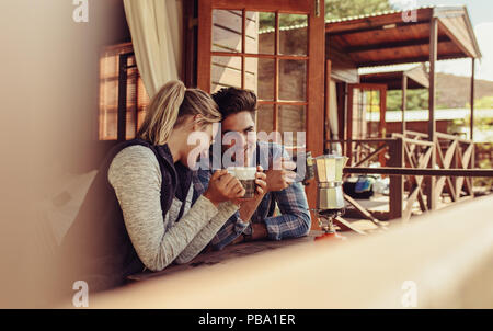 Loving young couple sitting outside a wooden cabin having coffee and relaxing. Man and woman drinking coffee together. Stock Photo