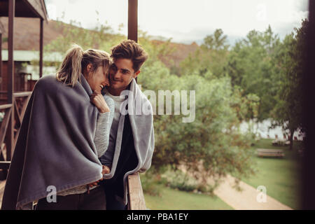 Loving man and woman standing in their hotel room balcony wrapped in blanket. Romantic couple in a blanket standing together on a winter holiday. Stock Photo