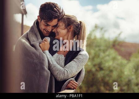 Young man embracing his girlfriend standing in a balcony, with both wrapped in blanket. Couple on winter holiday embracing each other and smiling. Stock Photo