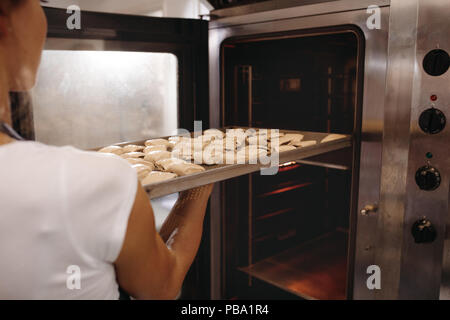 Female baker putting a baking tray full of rolls in oven. Woman baking rolls in a bakery. Stock Photo