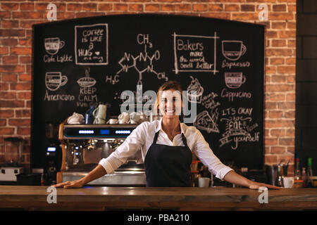 Portrait of confident female barista standing behind counter. Woman cafe owner in apron looking at camera and smiling. Stock Photo