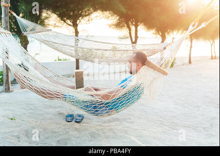 A man with a beard lying in a hammock with a laptop. Remote work on the beach Stock Photo