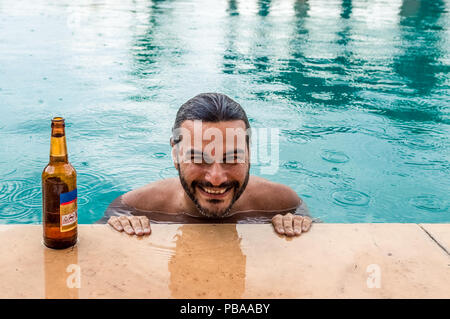 Happy brunet man smiling with a bottle of beer in a swimming pool while raining Stock Photo