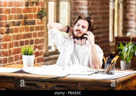 Bearded awesome male architect in white shirt with pencils and blueprint using mobile phone in loft office Stock Photo