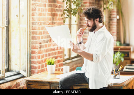 Young ambitious bearded architect in white shirt analyzing the details of his project in loft office Stock Photo