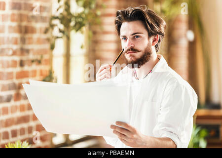 Bearded male architect in white shirt thinking and analyzing the details of his project in loft office Stock Photo