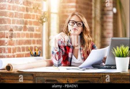 Cute ambitious female architect in plaid shirt wearing eyeglasses thinking about a new project in loft office Stock Photo