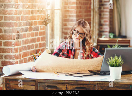 Happy attractive female architect in plaid shirt and glasses sitting and working with laptop in a loft office Stock Photo