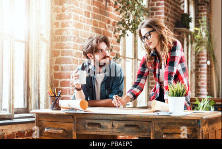 Female ambitious architect in glasses showing an architectural plan to her young bearded co-worker at the meeting in a loft sunny office Stock Photo