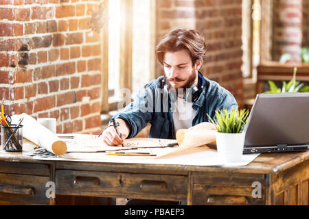 Concentrated young architect working with blueprint while sitting with laptop in a loft sunny office Stock Photo