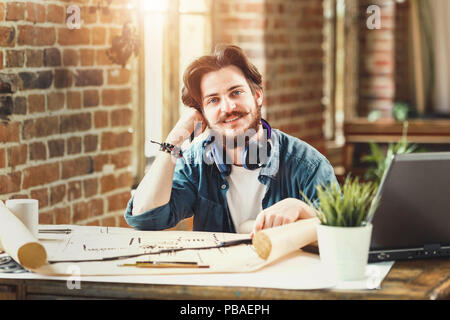 Male bearded architect smiling to the camera sitting at his desk at the office working on building plans copyspace creativity engineering project developing worker occupation career successful entrepreneur. Stock Photo