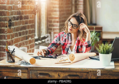 Young caucasian pretty building designer in glasses sitting alone at a table in a loft modern office and using tablet Stock Photo