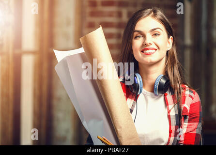Cute young long-hair female architect with headphones holding blueprints and smiling near the window in the loft modern office Stock Photo