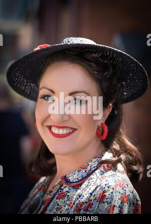 Close-up, front view portrait of smiling, pretty lady looking straight at camera in hat & floral frock, isolated at UK 1940's WWII event, in sunshine. Stock Photo