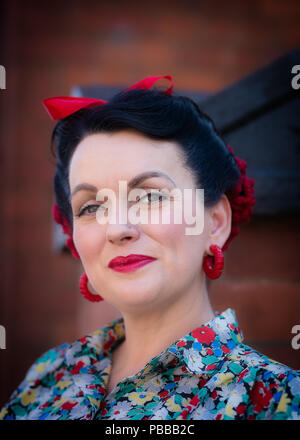 Close-up, front view portrait of attractive smiling lady looking straight at camera, red bow in hair & floral frock, isolated at UK 1940's WWII event. Stock Photo