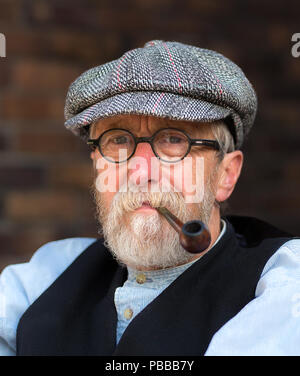 Close-up, front view portrait of senior man, bespectacled with pipe in mouth, wearing flat cap at Black Country Living Museum WWII 1940's event. Stock Photo