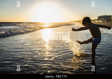 Baby boy playing makes splashes at sunset. Spanish southwest beach. He is kicking the waves Stock Photo