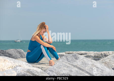 fit sports woman exhausted after exercise sitting on rocks by the sea with her head in her hands Stock Photo
