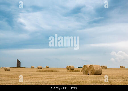 East Lothian, Scotland, United Kingdom, 28th July 2018. UK Weather: Dark clouds gather with a stormy threatening sky over recently harvested grass field with round hay bales and a stormy sky over a ruined windmill in the distance Stock Photo