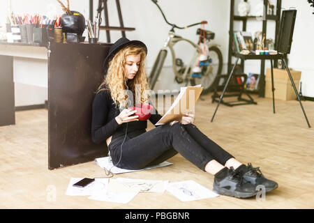 Blonde long hair curly feemale artist in black hat and black clothes drinking coffee and looking on the sketch, she sitting on the floor Stock Photo