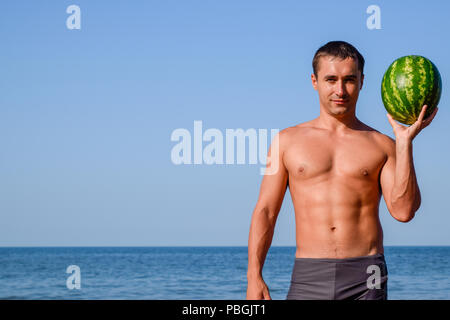 A sporty man is standing on the beach and holding a whole watermelon. A ripe watermelon in the hands of a man. Stock Photo