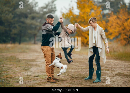 Happy parents have fun and lift up their daughter at forest path next to jumping dog during walk in autumn forest. Stock Photo