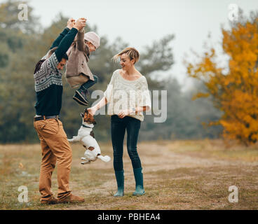 Happy parents have fun and lift up their daughter at forest path next to jumping dog during walk in autumn forest.