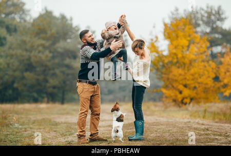 Happy parents have fun and lift up their daughter at forest path next to jumping dog during walk in autumn forest.