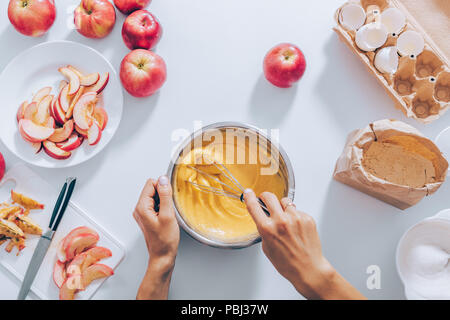 Top view of female hands holding whisk and mixing bowl with batter for apple pie. Flat lay arrangement woman preparing autumn fruit cake on table with Stock Photo