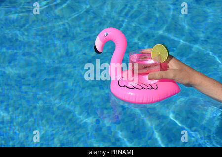 Summer vibes! Woman’s hand, with a cold drink in an inflatable pink flamingo floating drinks holder in a swimming pool Stock Photo