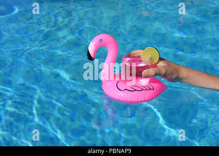 Summer vibes! Woman’s hand, with a cold drink in an inflatable pink flamingo floating drinks holder in a swimming pool Stock Photo