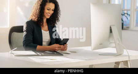 Smiling woman entrepreneur at her desk in office checking her mobile phone. Woman sitting in front of computer looking at cell phone. Stock Photo