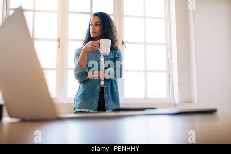 Woman entrepreneur standing in office holding a coffee cup looking away. Woman standing near her table drinking coffee with a laptop in the foreground Stock Photo