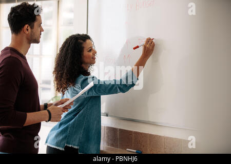 Office colleagues discussing monthly budget and plans on a whiteboard. Businesswoman writing on the board while her colleague holds a paper in hand. Stock Photo