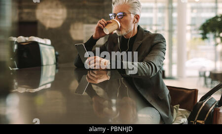 Mid adult businessman sitting at a cafe drinking coffee and using smart phone. Caucasian man in earphones looking at his cellphone in coffee shop. Stock Photo