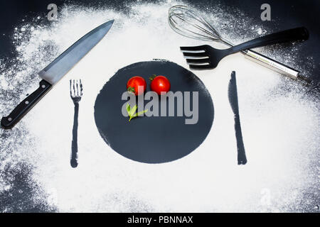 on a black marble background flour powder powdered snow silhouette plate, fork, knife appliances in the center lies a round slice of tomato and mint Stock Photo