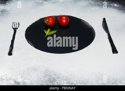 on a black marble background flour powder powdered snow silhouette plate plate fork knife appliances in the center tomato and mint Stock Photo