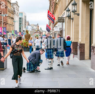 People dressed for wedding with men in kilts stopping to tie shoelace and young woman turning head, Buchanan Street, Glasgow, Scotland, UK Stock Photo